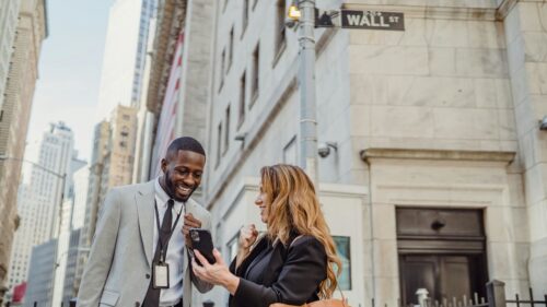 Two business professionals in suits smile and look at a phone on a bustling city street under a Wall St sign, embodying the harmony of finance and tech.