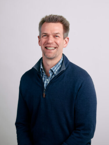 A smiling man in a blue sweater over a checked shirt stands against a plain white background, embodying the principles of responsible tech with his forward-thinking approach.