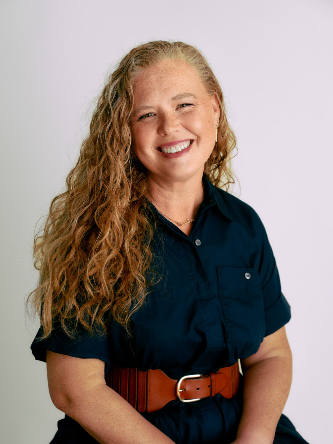 A smiling woman with long curly hair, wearing a dark shirt and brown belt, is seated against a plain light background, embodying the harmony between technology and human spirit.