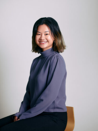 A smiling person with shoulder-length hair, wearing a purple blouse, is seated against a plain background.